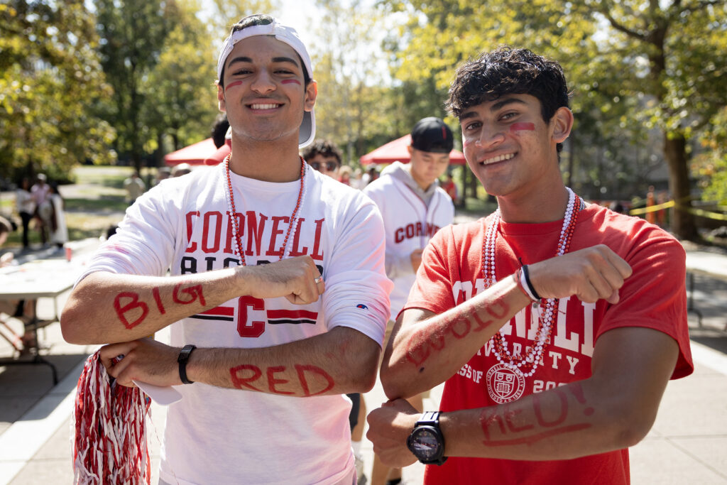 Two students smile while showing off paint on their arms that says "Go Big Red," facepaint, Cornell shirts, and red and white beaded necklaces.