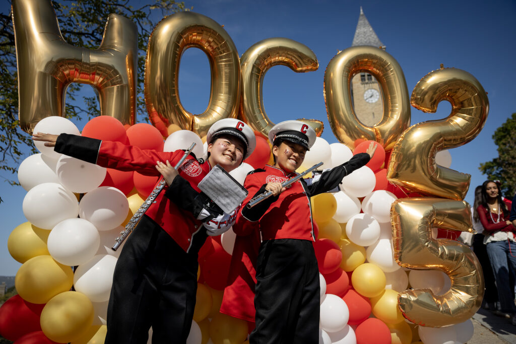 Two flute players in the Big Red Band pose with their hands in the air in front of a large balloon installation that says "HOCO25."