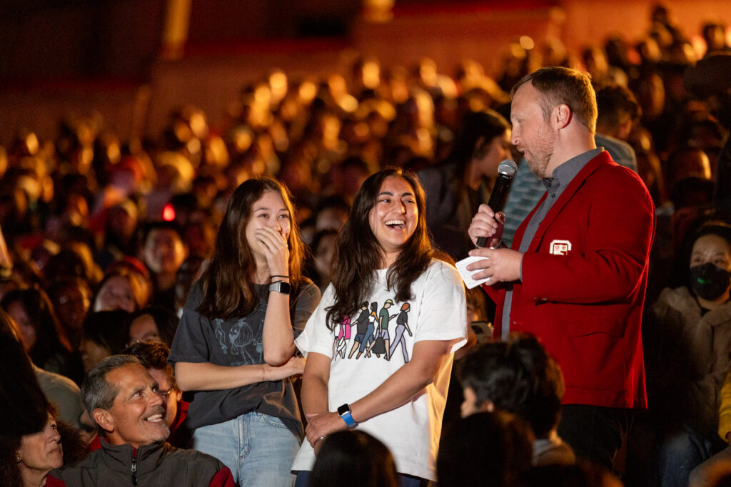 Corey Ryan Earle stands and holds a microphone while talked to two students during a crowded homecoming event.