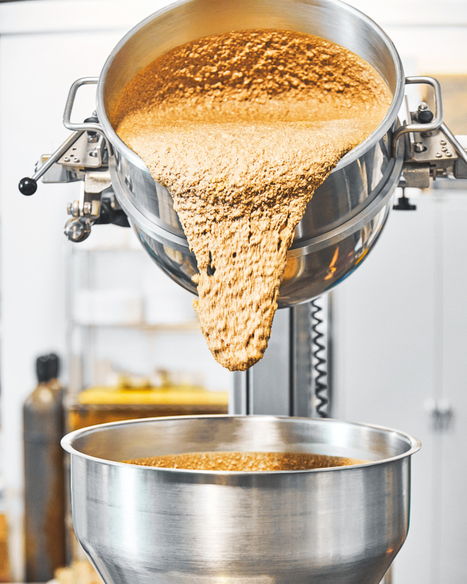 Fresh nut butter being poured from one metal industrial container into another.