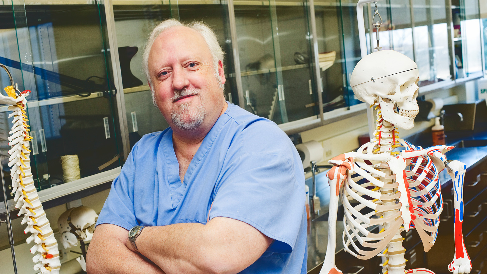 Jim Perkins smiles next to a teaching skeleton in an anatomy classroom.