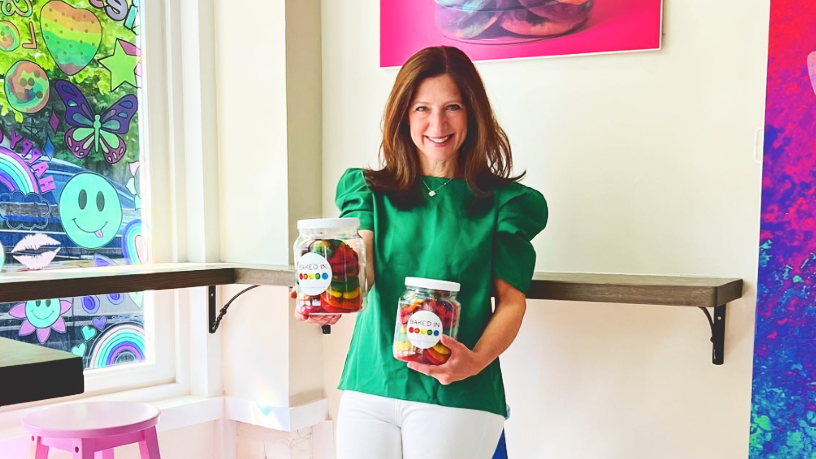 Julie Waxman stands in her colorful shop and holds two jars of rainbow cookies up while smiling.