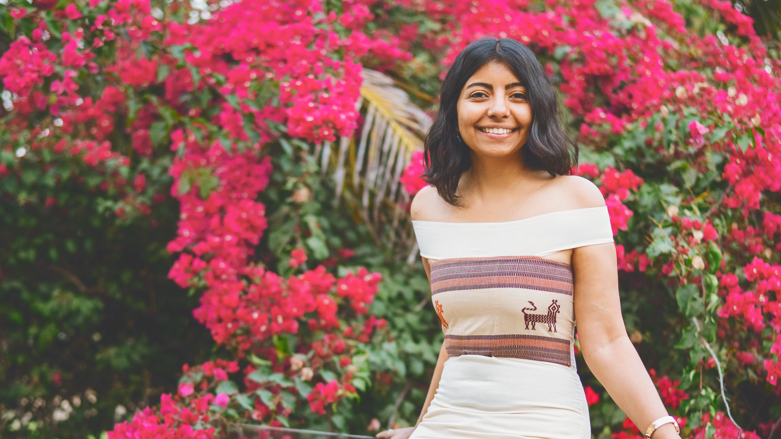 Aishwarya Lahariya smiles standing in front of pink flowers and wears a white and brown off the shoulder dress from Jiwya.