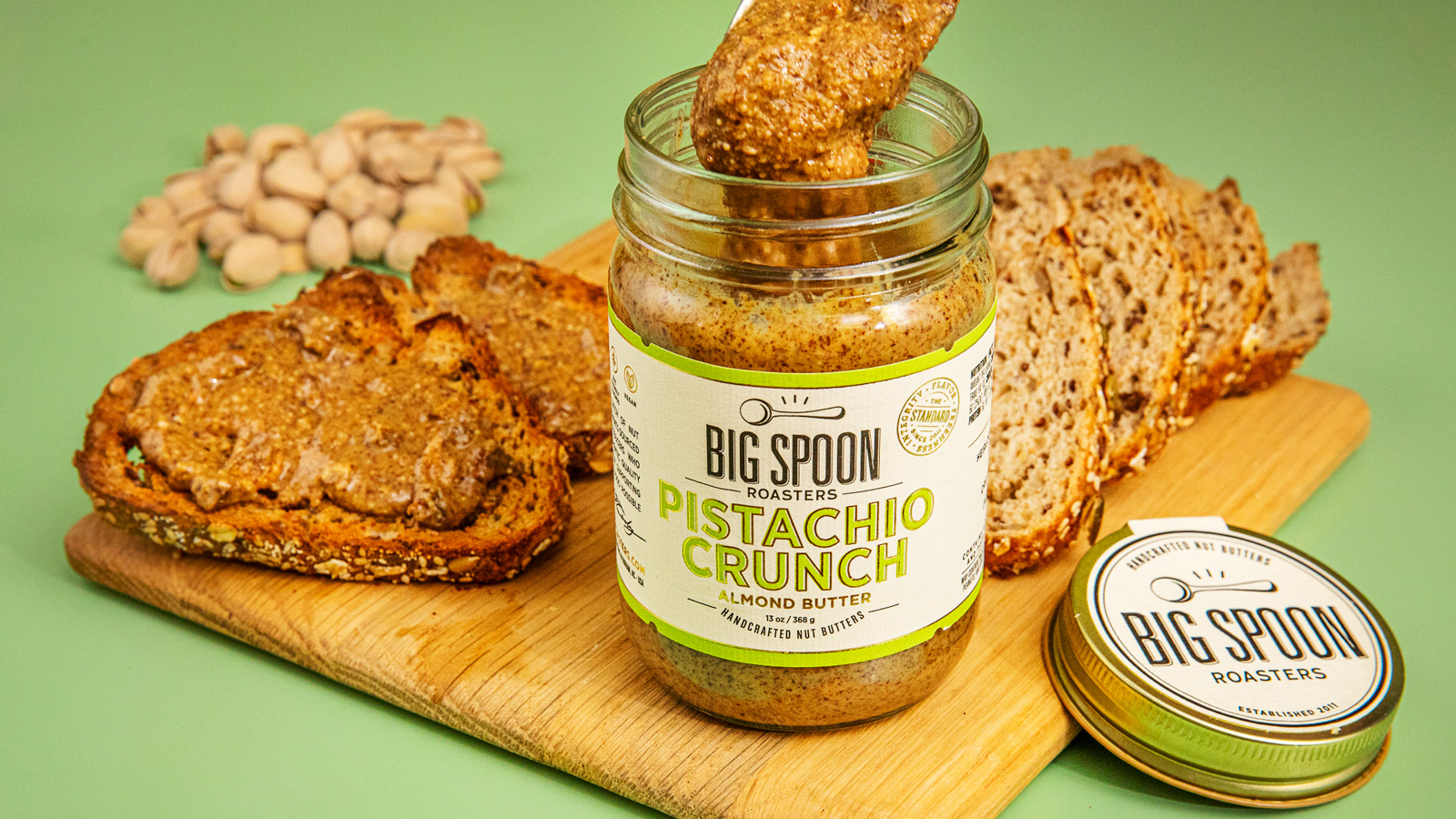 An open jar of pistachio crunch almond butter with toast spread with the butter on a wood cutting board.