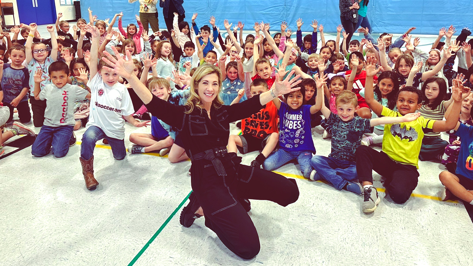 Heather Alexander smiles with her hands in the air kneeling in front of a crowd of smiling and waving kids.