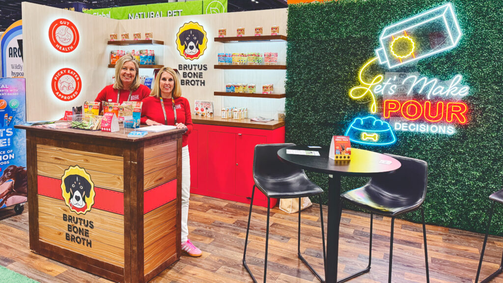 Kim Hehir with her sister at a food trade show featuring Brutus Bone Broth products on shelves behind them and a light-up sign that says "Let's make pour decisions."