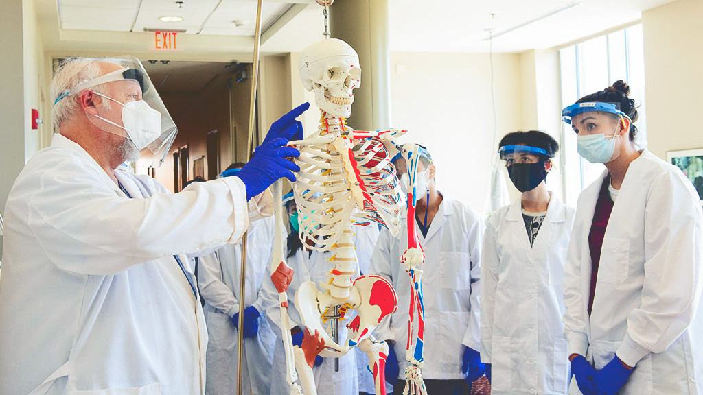 Jim Perkins and a group of his students wear white lab coats, gloves, and face masks while inspecting a teaching skeleton.