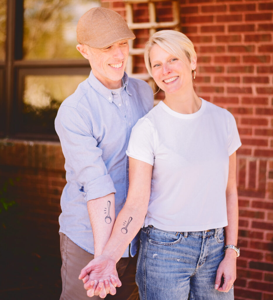 Megan and Mark Overbay hold hands and smile while showing their matching spoon tattoos on their wrists.