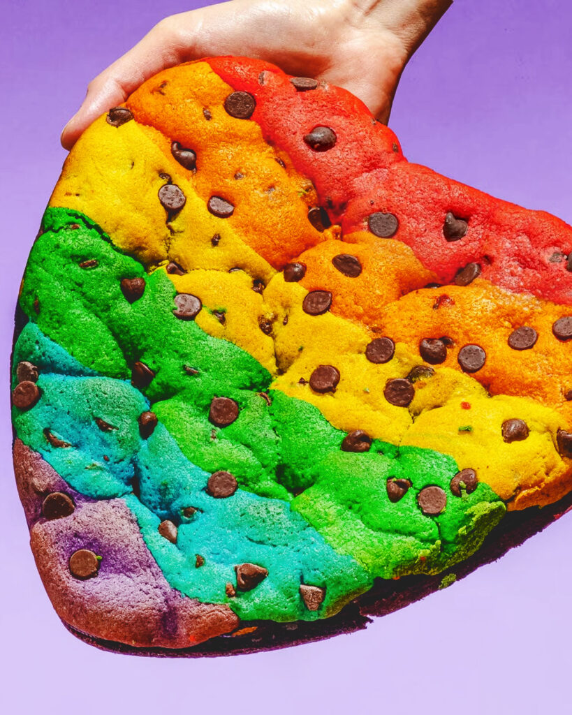 Hands hold a rainbow striped heart brookie cake for pride month.