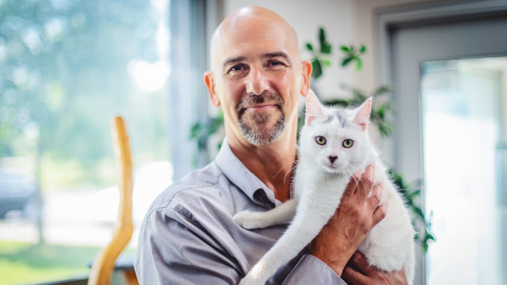 Bruce Kornreich smiles while holding a white cat in his arms.