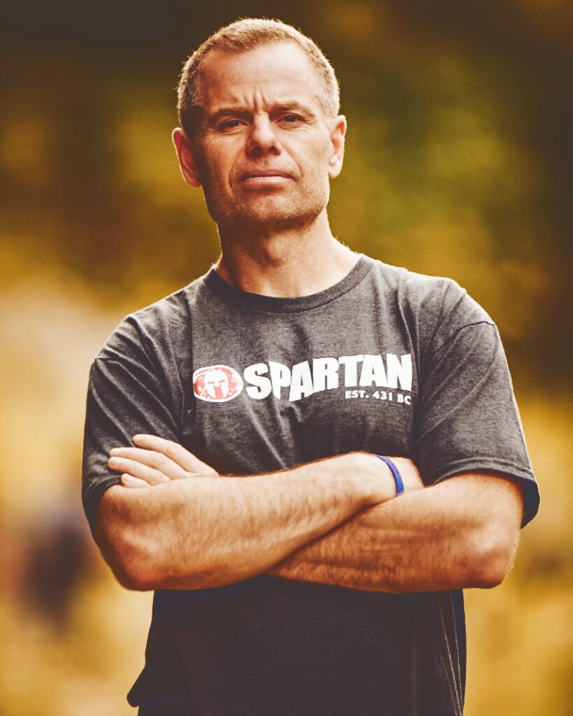 Joe De Sena with his arms crossed in front of him wearing a Spartan shirt.
