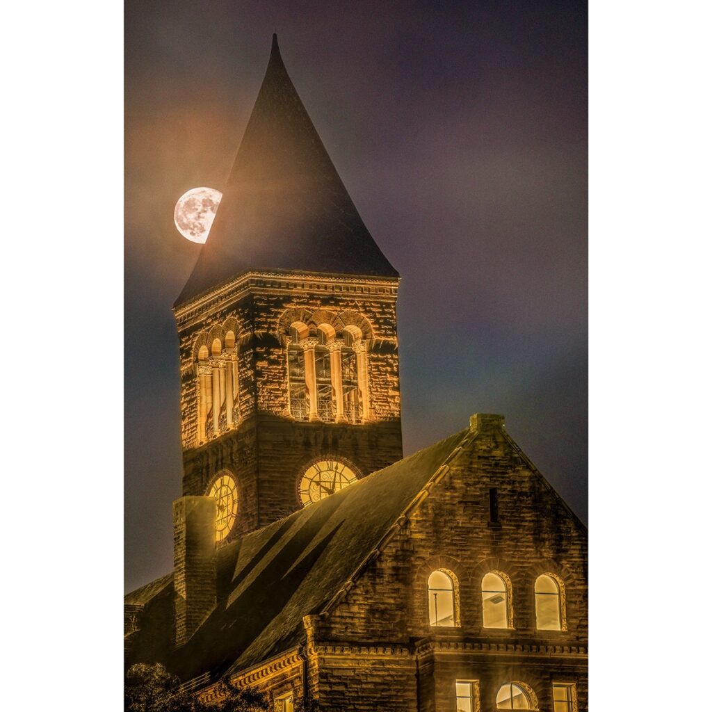 A photo of McGraw Tower lit up at night with the moon behind it.