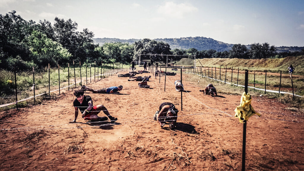 Participants crawl in the dirt underneath barbed wire.