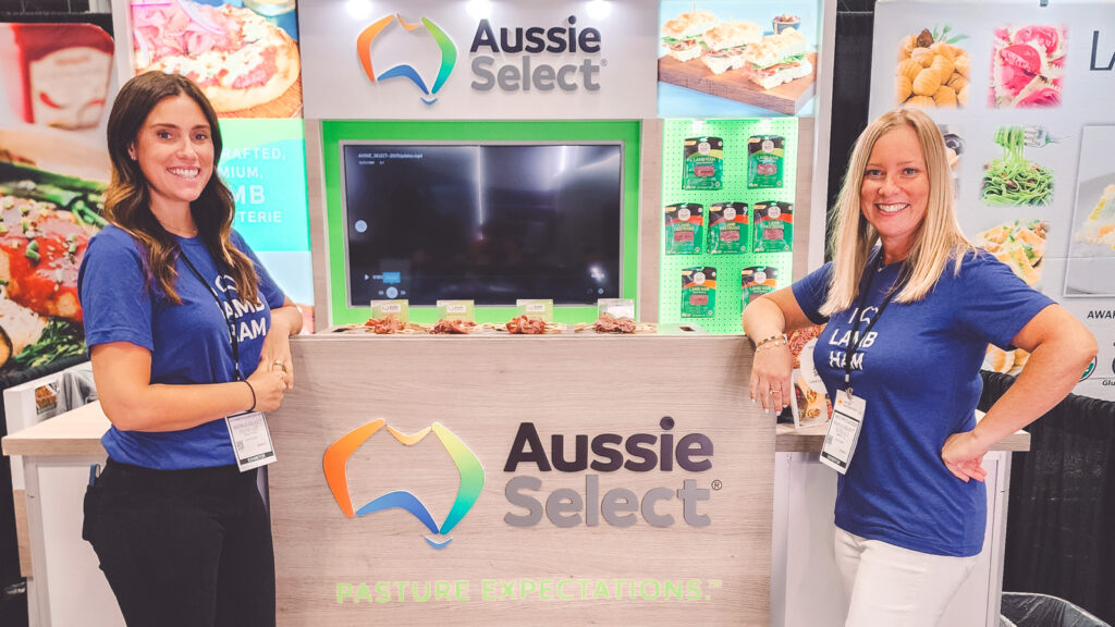 Jaclyn Oyola and a coworker smile in front of a Aussie Select stand with samples at a food show.
