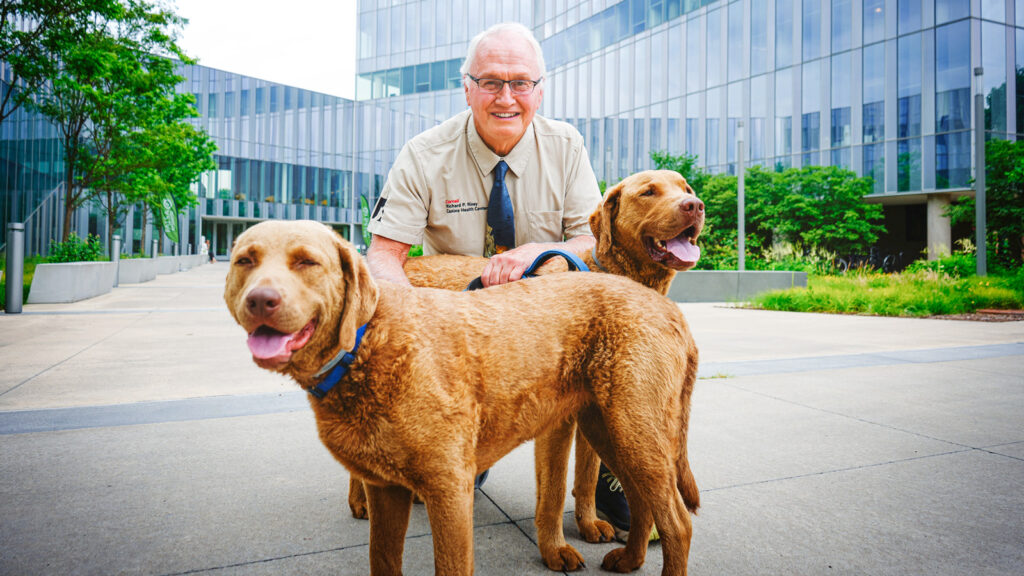 Rory Todhunter kneels and smiles with two dogs in front of the College of Veterinary Medicine.