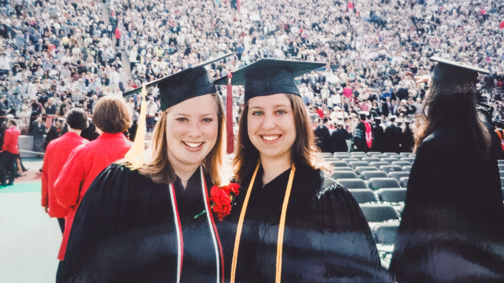 Jaclyn Oyola and a friend at graduation in their regalia with a crowd of people behind them.