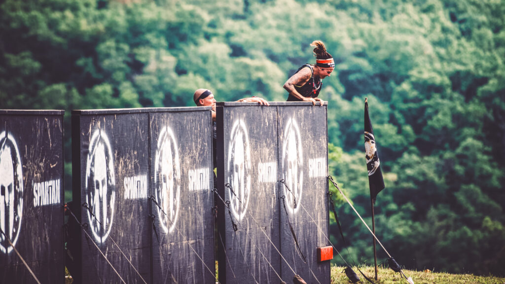 Two competitors hoist themselves over a wall mid Spartan race.