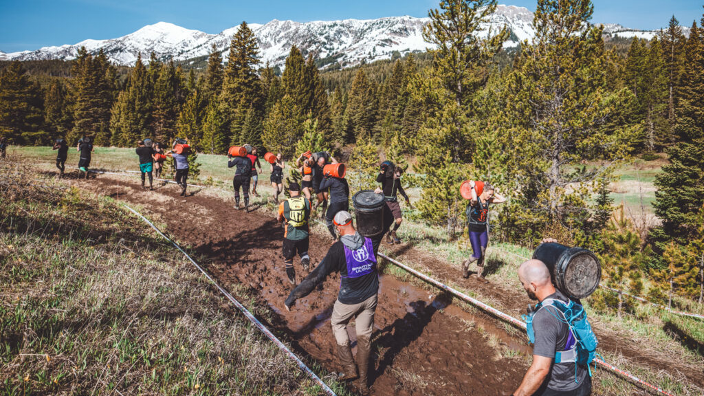 Participants carry weighted barrels over one shoulder while competing in the Spartan race with trees and mountains in the background.