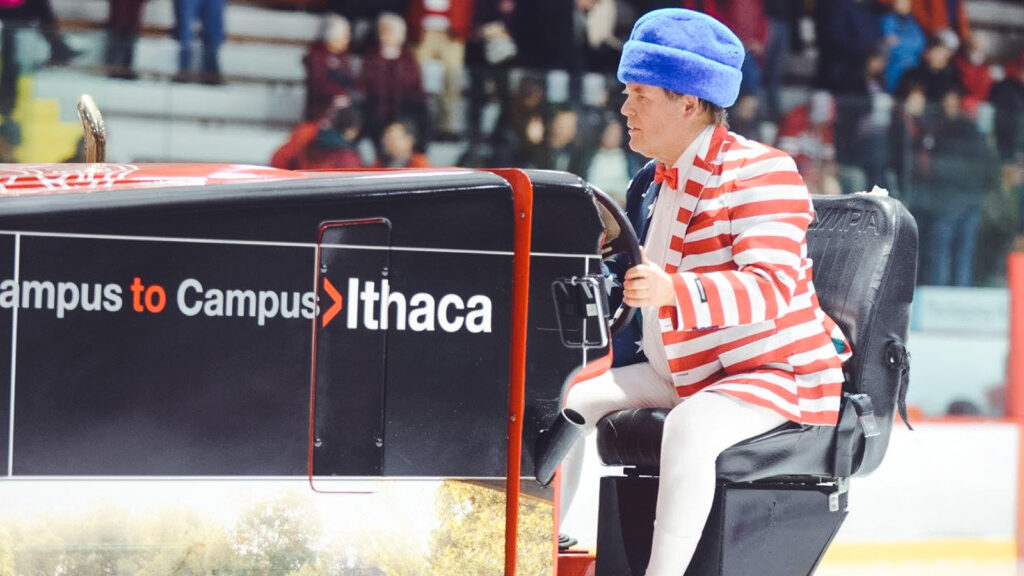 Dave Nulle driving an ice resurfacer at Lynah Rink, dressed in a red striped jacket, white tights and a blue hat