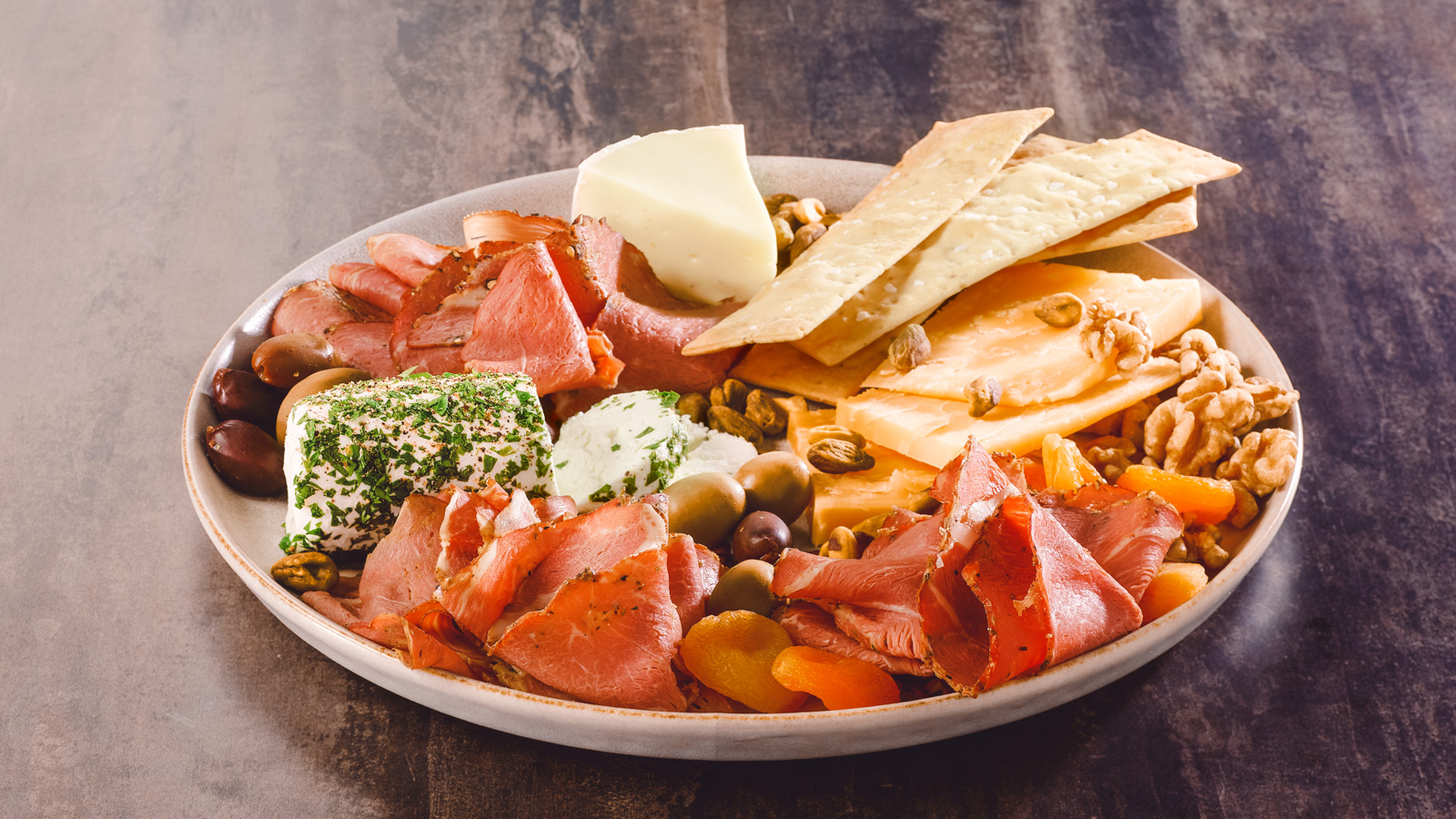 A charcuterie board with sliced lamb, cheeses, nuts, dried fruit, and olives.