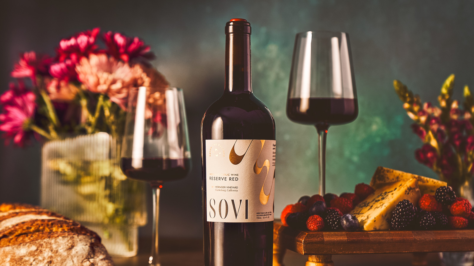 A bottle od Sovi reserve red with two glasses poured with a cheese and berry tray behind it.
