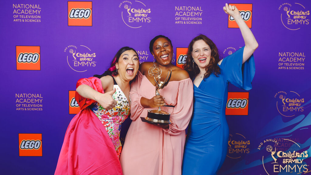 Monique Hall smiles and holds her Emmy award on the red carpet with two colleagues.