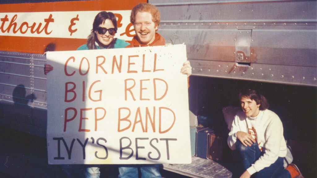 Heather Doering Mitchell and Scott Pesner hold a "Cornell Big Red Pep Band: Ivy's Best" sign in front of a bus in 1987