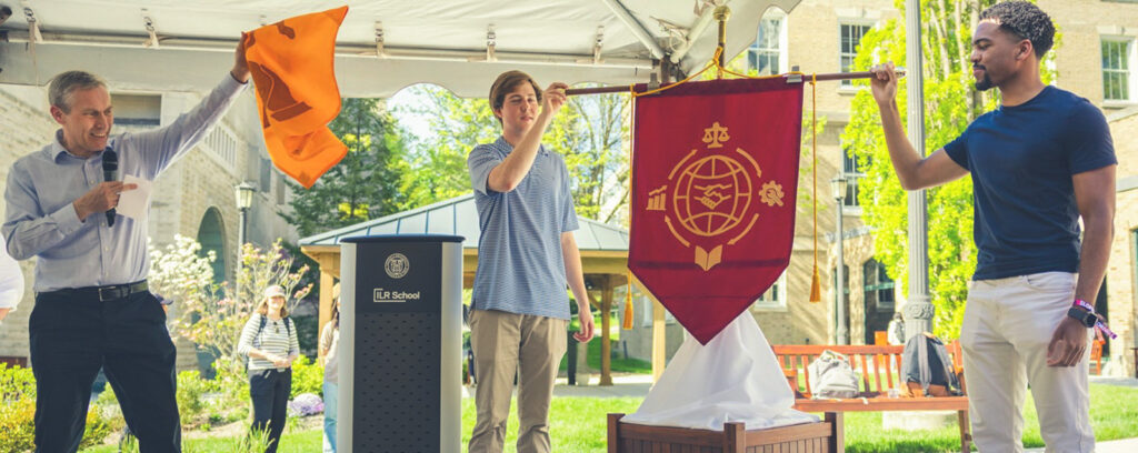 ILR Dean Alex Colvin, Ph.D. '99, unveils the school’s new ceremonial banner in May 2024. Holding the updated flag are Patrick Raczka ’25 (left) and Tyler Bonaparte ’25 (right), who were part of the student team that created the winning redesign.