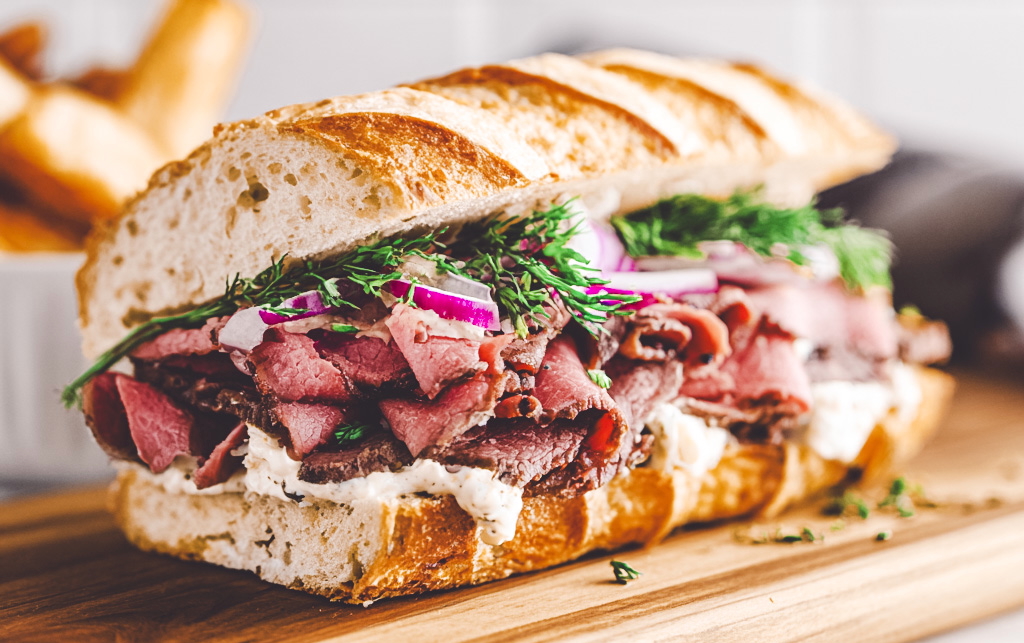 A lamb pastrami sandwich sits on a wooden cutting board in a kitchen.