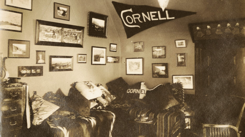 a Cornell dorm room in 1906 includes a Cornell pennant on the wall and a Cornell pillow on a bed
