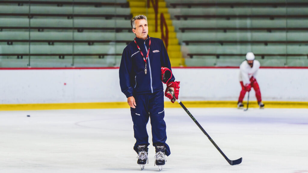 Doug Derraugh skates on the ice holding a hockey stick with a whistle around his neck during a practice.