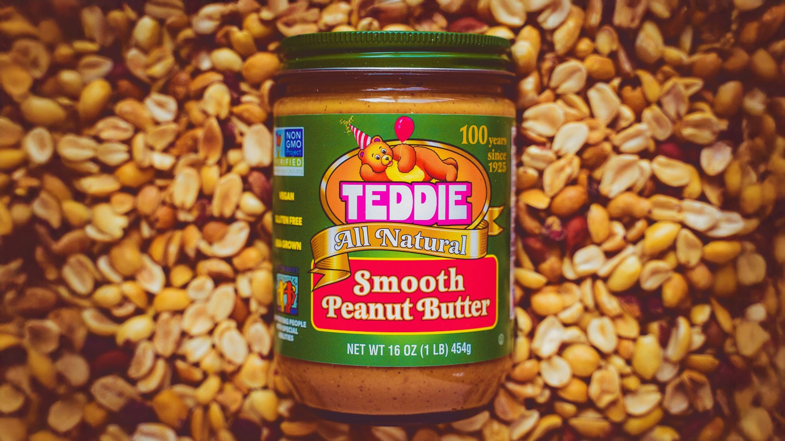 jar of Teddie All Natural Smooth Peanut Butter with 100th anniversary label design on a background of shelled peanuts