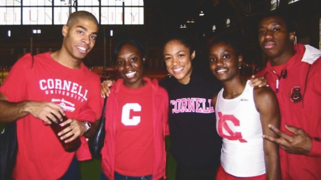 Troy Mullins smiles with four of her friends on the track and field team at Cornell inside Barton Hall, all of them wearing Cornell shirts.