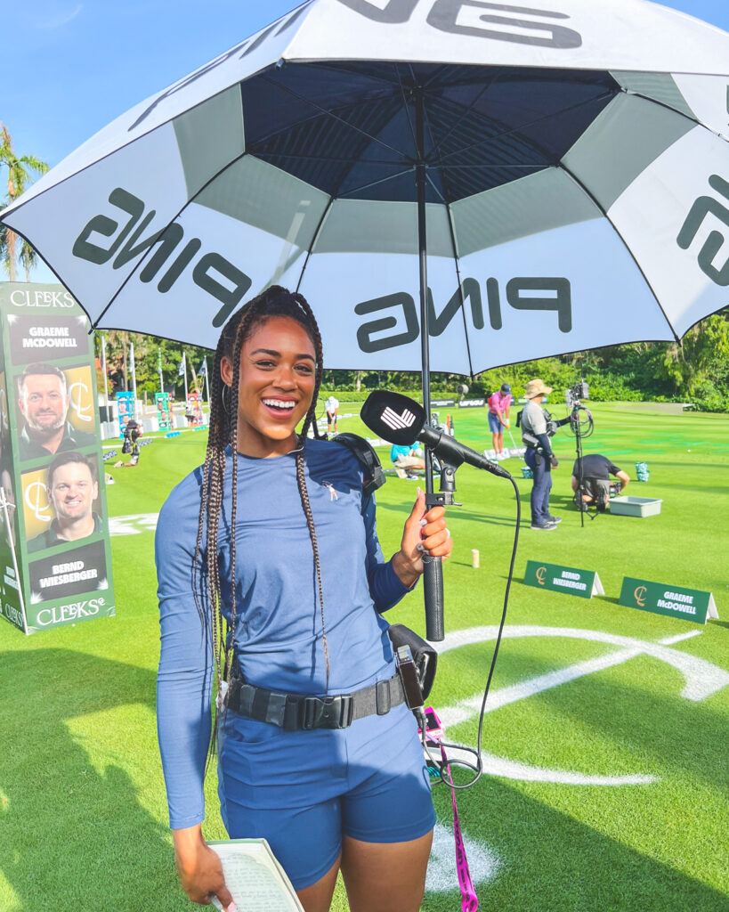 Troy Mullins smiles while holding an umbrella with a microphone attached to it during an on course reporting gig at a golf tournament.
