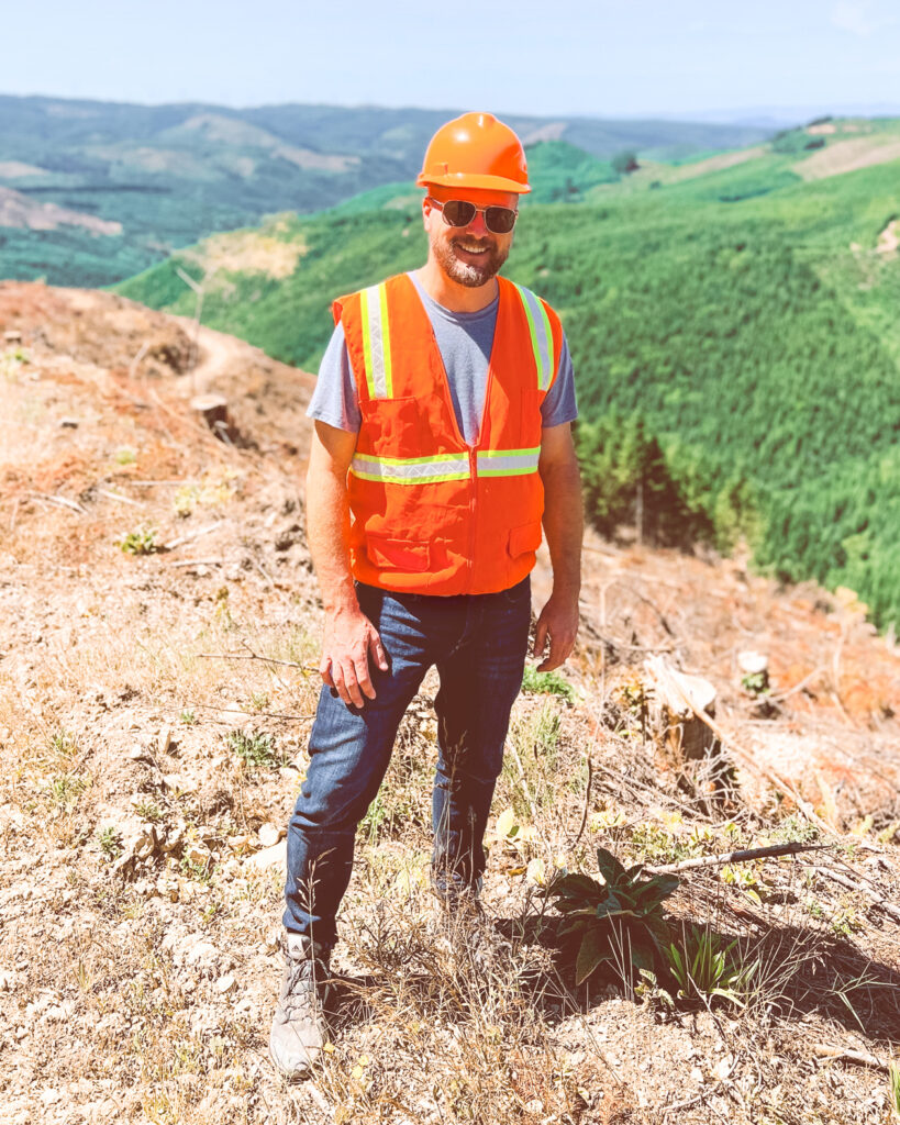 Trent Preszler wearing an orange vest and hard hat at a logging site with forests in the background