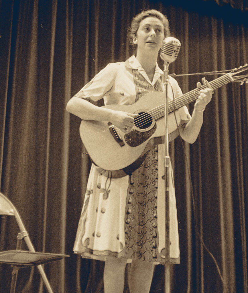Ellen Stekert wearing a dress on stage at Cornell signing and playing the guitar in 1960.