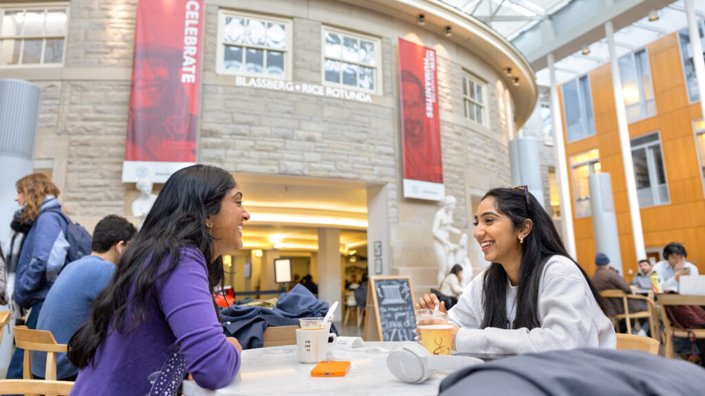 Two students sit at a table in Klarman Hall atrium smiling and talking to each other with coffee and food