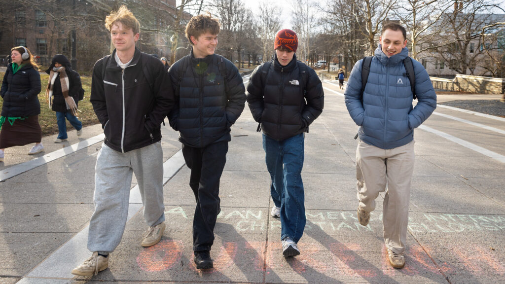 Four students wearing jackets and backpacks walk in a row together on Ho Plaza on the first day of classses