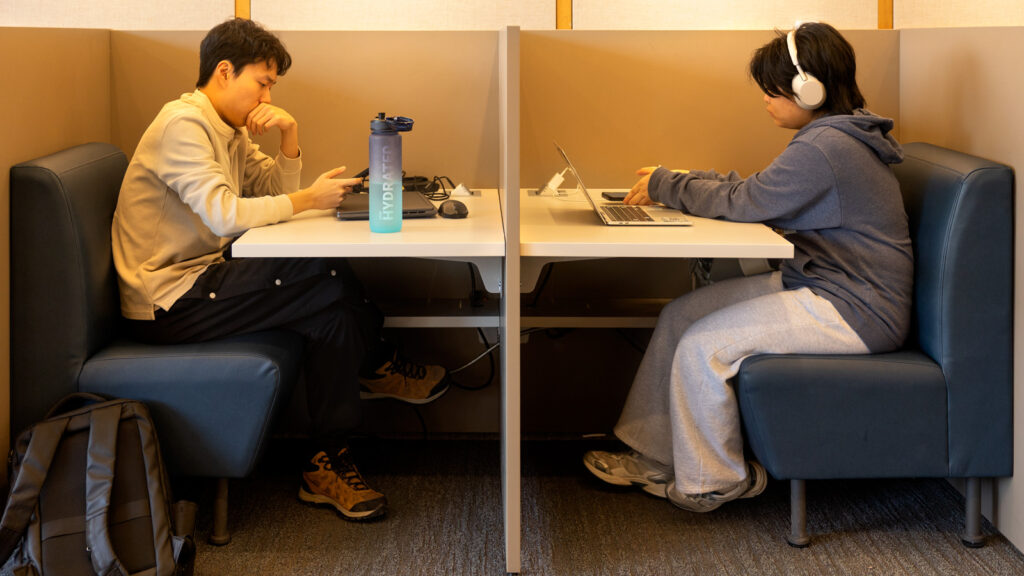 Two students sit at booths in Olin Library with their laptops and phones. One student wears a pair of headphones.