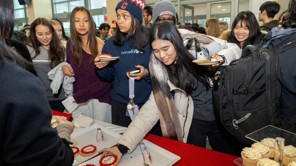 Students stand in front of a table with plates and pick up Cornell Duffield Engineering school branded cookies.