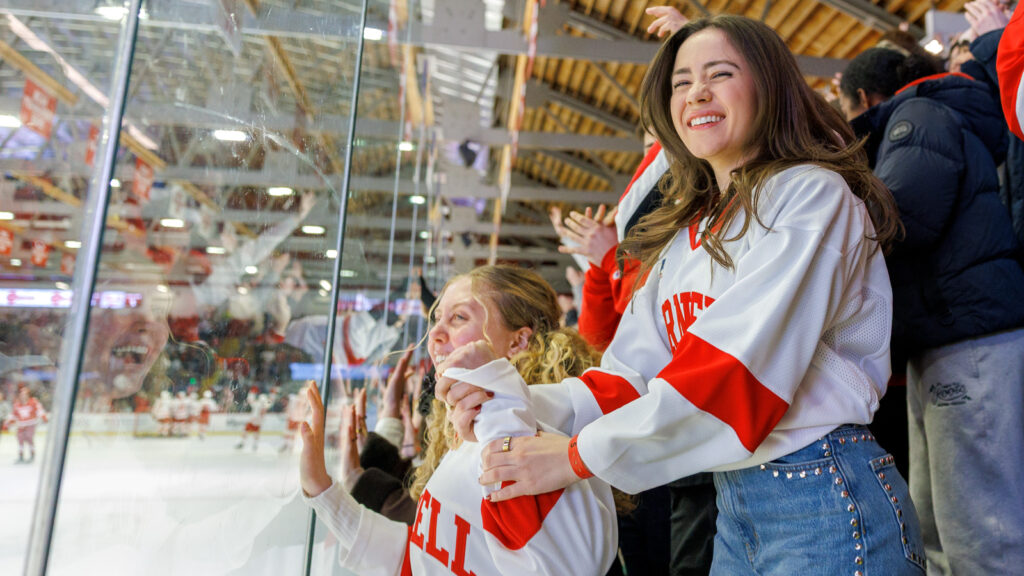 Fans cheer in the stands and put their hands to the glass wearing Cornell apparel.