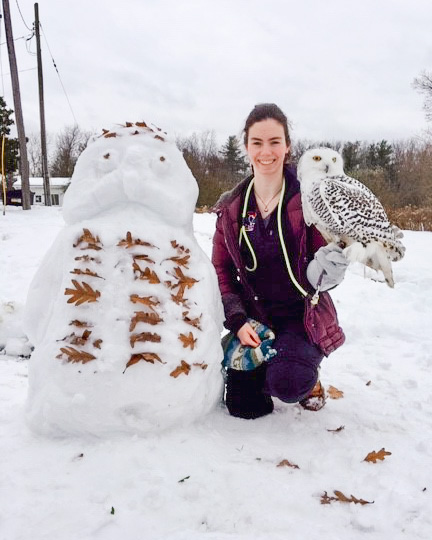 A student handler outside in the snow with a snow sculpture of an owl while Denali, a snowy owl, sits on her glove.