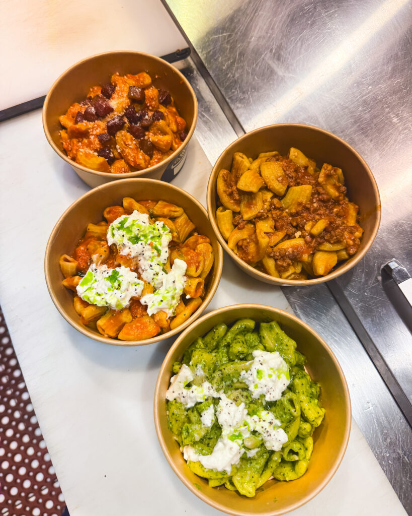 Four to-go bowls of rigatoni pasta with varying sauces, including bolognese and pesto, on them.