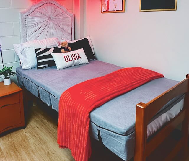 A dorm room featuring the Expand A Bed mattress topper on the bed and decor in the room including light up sign and posters.
