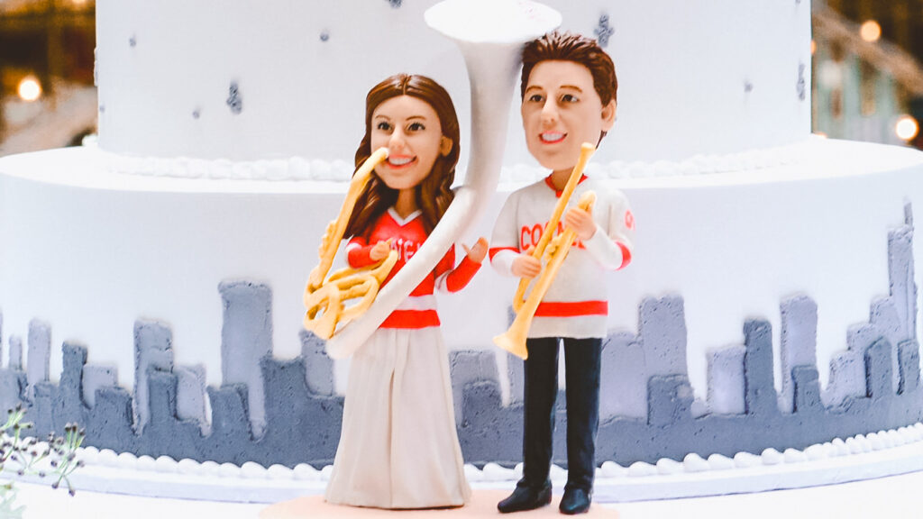 A cake topper dpiecting a couple in Big Red hockey shirts holding musical instruments