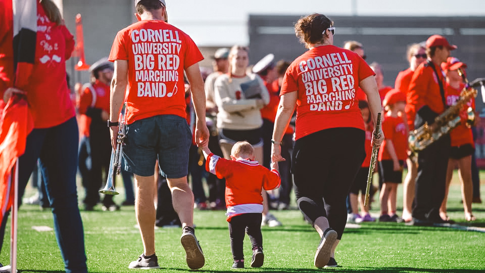 A couple wearing Big Red Band t-shirts walking away holding a toddler's hands, at Homecoming
