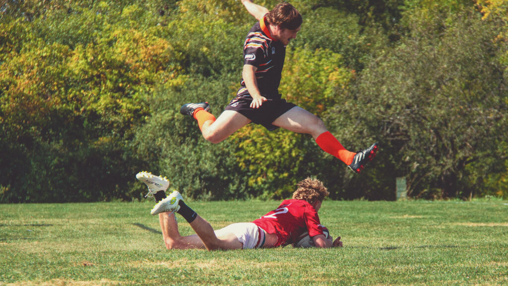 A Cornell rugby player dives onto the ground with the ball while an opponent hurdles over him in the air.