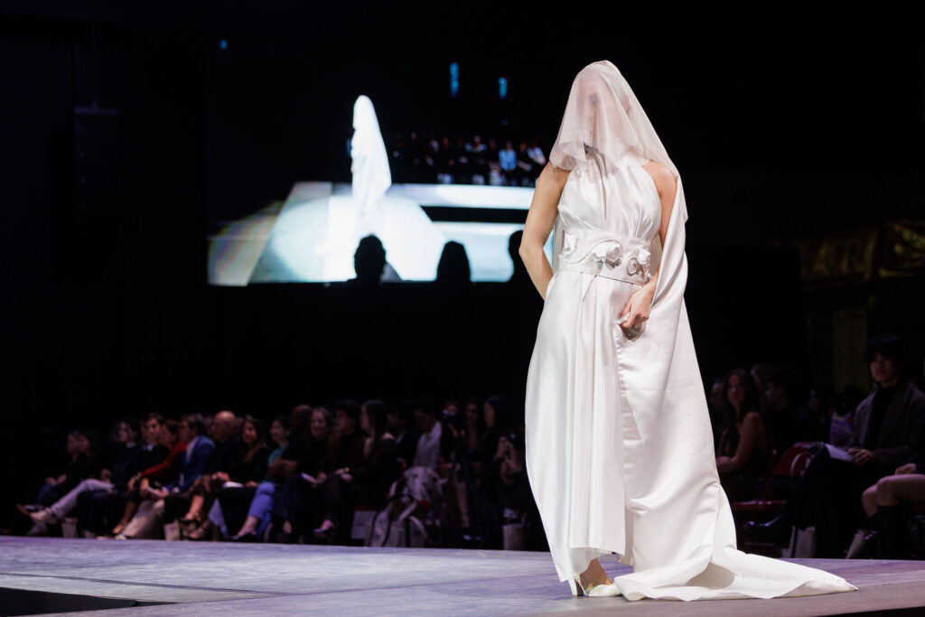 A model stands at the end of the runway in wearing a long white silky dress with a veil covering her face