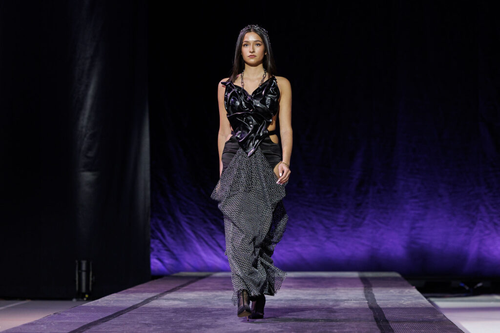 A student walks down the runway wearing a black outfit featuring several different materials.