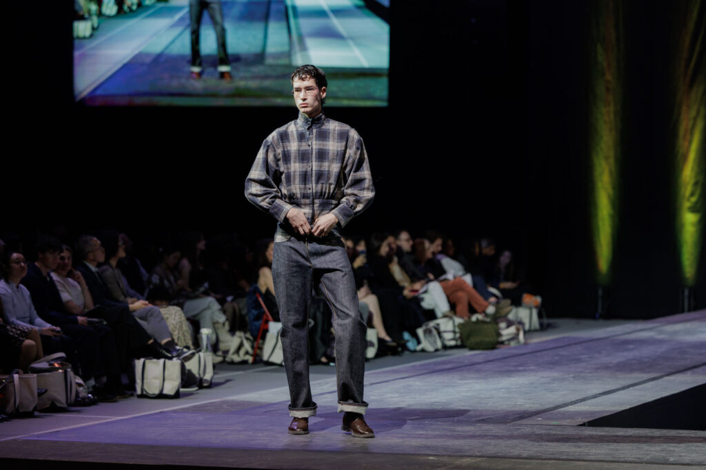 A model poses at the end of the runway in a flannel that is cinched at the waist and jeans.